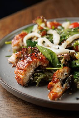 Izgara balık, yeşillik ve taze sebzelerin yer aldığı bir tabak dolusu yemek yuvarlak gri bir tabakta servis edilir. Düzenlemede renkli malzemeler ve farklı dokular vurgulanıyor.