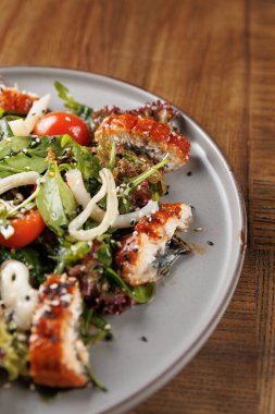 Gri bir tabakta ızgara et, taze yeşiller, kiraz domatesleri ve otlardan oluşan renkli bir salata sergilenmektedir. Ahşap masa arkaplanlı kırsal bir ortam..