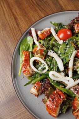 Taze bir salata ızgara balık, kiraz domatesleri, yeşil fasulye, soğan ve karışık sebzelerle sunulur. Tahta bir masada yuvarlak bir tabakta servis edilir..