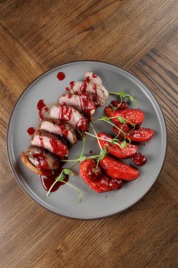 Bir tabak taze meyvenin yanında kırmızı soslu dilimlenmiş et içerir. Tabak ahşap bir masanın üzerinde, renklerin ve dokuların bir karışımını gösteriyor..