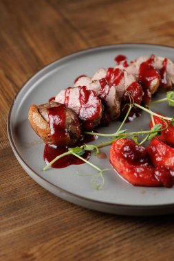 Pişmiş et dilimleri gri bir tabakta düzenlenir. Kırmızı bir sos etin üzerine serpiştirildi. Taze domates dilimleri ve yeşil garnitür ahşap bir masada etin yanına konur..