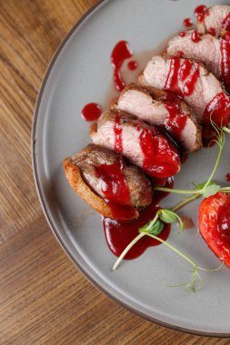 Dilimlenmiş et parçaları, canlı sosla kaplanmış, garnitürlerin yanında yuvarlak bir tabakta oturur. Ahşap masa yemek sırasında yemek sunumunu güçlendirir..