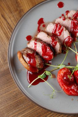 Dilimlenmiş et parçaları üzerine kırmızı sos serpiştirilmiş bir tabağa konuyor ve yanında süslemeler getiriliyor. Sunumda bir yemek alanında yemek sergileniyor..