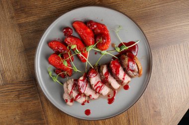 Dilimlenmiş ördek göğsü, üzerine çilek sosu serpilmiş gri bir tabakta güzelce düzenlenir. Taze kırmızı meyve ahşap bir masada tabağa eşlik eder..