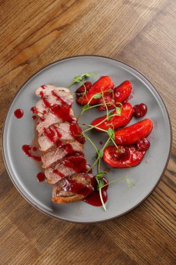 Dilimlenmiş bir tabak çilek soslu ördek göğsüne kızarmış vişneli domates eşlik eder. Yemek ahşap bir masada güzelce düzenlenmiş..