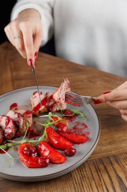 Bir el, ahşap bir masada sunulan böğürtlen sosuyla servis edilen et tabağını keserken bir çatal ve bıçak tutar. Yemek zarif bir şekilde sunuluyor ve canlı renkler sergileniyor..