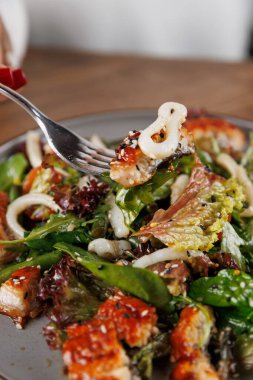 Bir insan bir parça ızgara tavuk ve salatayla çatal tutar. Tabakta yapraklı yeşiller, sebzeler ve bir sos serpintisi yer alıyor. Rahat bir atmosferde öğle yemeği için mükemmel..