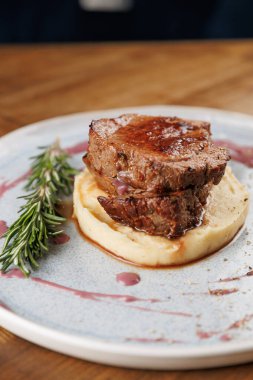 Bir dilim köfte kremalı patates püresinin üzerine serpilmiş sosla dinleniyor. Taze biberiye, çağdaş bir ortamda dekoratif bir tabakta sunulan yemeğe tat katar..