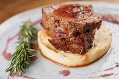 Yumuşak pişirilmiş, yumuşak patates püresinin üzerine yerleştirilmiş, biberiye filizi ile süslenmiş, doyurucu bir biftek. Yemek şık bir tabakta, lezzetleri vurguluyor..