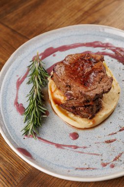 Kremalı patates püresi üzerine ızgara sığır eti. Taze biberiyeyle süslenmiş ve böğürtlen sosuyla sulandırılmış. Tabak ahşap bir masada yuvarlak mavi bir tabakta servis edilir..
