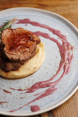 Sığır filetosu pürüzsüz patates püresi yatağında, mavi bir tabakta çilek sosuyla süslenmiş. Kırsal bir ahşap masa yemek için sıcak bir atmosfer sağlar..