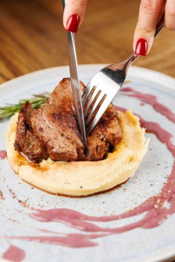 Bir insan ızgara eti bir tabakta kesiyor. Et, kremalı patates püresinin üzerine konur. Renkli bir sos tabağı sarar ve sunumunu geliştirir..