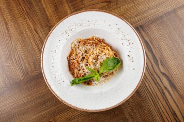 Üzerine et sosu ve peynir serpiştirilmiş spagetti dolu bir tabak ahşap bir masaya yerleştirilir. Taze fesleğen yeşil bir dokunuş ekler..