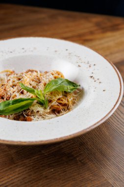 Et soslu spagetti servisi, rendelenmiş peynir ve taze fesleğen yaprakları yuvarlak beyaz bir tabakta duruyor. Ahşap masa yemek deneyimini arttırır..