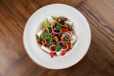 Renkli bir salata yuvarlak beyaz bir kasede servis edilir. Tabakta marul, soğan, kırmızı biber, tavuk ve fındık yer alıyor. Tahta bir masaya konur..