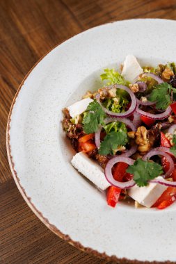 Taze bir salatada kırmızı dolma biber, soğan ve ceviz ve bitkilerle karıştırılmış yeşil marul da dahil olmak üzere doğranmış sebzeler yer alıyor. Tahta bir masada beyaz bir tabakta servis edilmiş..