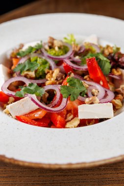 Canlı bir salatada kırmızı biber, feta peyniri, ceviz ve dilimlenmiş kırmızı soğan bulunur. Taze otlar tat katar. Tabak ahşap bir masada sunuluyor, Ukrayna mutfağı sergileniyor..