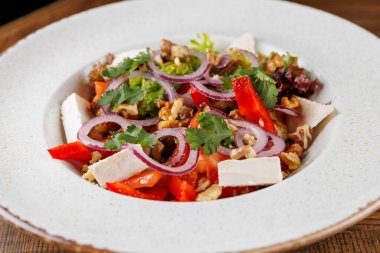 Canlı bir salata beyaz bir tabakta servis edilir. Domates ve biber gibi taze sebzeler içeriyor, üzerine soğan, bitki ve fındık konmuş, dokuma ve tat karışımı sunuyor..