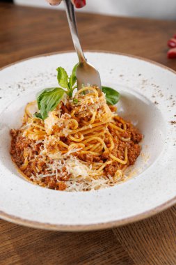 Birisi beyaz tabakta peynirli et soslu makarna döndürmek için çatal kullanıyor. Ahşap masa bir restorandaki yemek deneyimine sıcaklık katar..