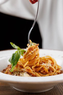 Çatal, bir spagetti spiralini beyaz bir kaseden kaldırır. Tabakta erimiş peynir, taze fesleğen ve ahşap bir masada servis ediliyor. Yemek sahnesi sıcak ve davetkar..