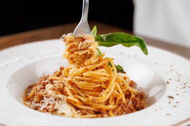 Et sosu ve taze fesleğenle kaplı bir çatal üzerinde bir spagetti servis edilir. Makarnanın üzerine beyaz bir tabakta rendelenmiş peynir serpiştirilir. Ayar yumuşak ışıklandırmayla davetkar görünüyor..