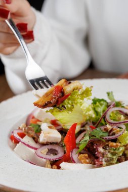 Bir insan beyaz kaseden salatanın bir kısmını kaldırmak için çatal kullanır. Tabakta bir restorandaki ahşap masada yeşiller, salatalıklar, domatesler ve tavuk yer alıyor..