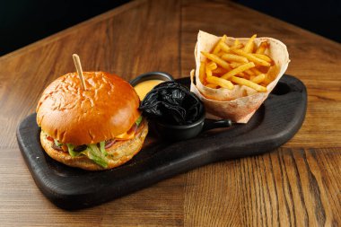 Sulu bir burger, tahta bir tahtanın üzerine konserve edilmiş altın patates kızartmasının yanına konur. Ayar, yemeğin cazibesini arttıran koyu ahşap tonlara sahip..