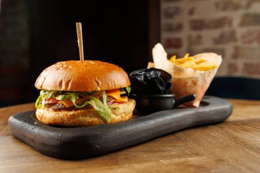 Tahtaya kızarmış patates ve sosun yanına bir hamburger konur. Ayarda sıcak bir ışık ve kırsal bir atmosfer yer alıyor. Yemekler hazır..