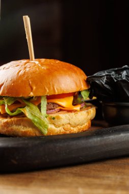 Bir hamburger ahşap bir tabakta oturur. Hamburgerde çörek, peynir, marul ve yanında siyah bir kabın içinde patates kızartması var. Ayar gündelik ve davetkar..