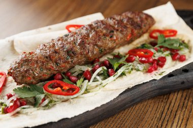 Düz ekmeğe konmuş kebap taze bitkiler, kırmızı biberler ve ahşap bir tabakta nar tohumları içerir. Sahne sıcak bir atmosferde lezzetli bir yemeğin altını çiziyor..