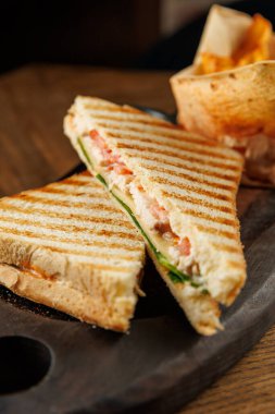 Taze sebze ve etle dolu ızgara bir sandviç ikiye bölünür ve katmanları ortaya çıkar. Tahta bir tabağın üzerinde çıtır çıtır cipsler oturuyor ve cazip bir yemek sahnesi yaratıyor..