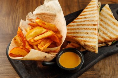 Bir yemek tabağı seçimi ahşap bir masada sergilenir. Sıcak bir sandviç, kızarmış patates ve küçük bir kase sosla servis edilir. Lezzetli bir yemek deneyimi yaratır..