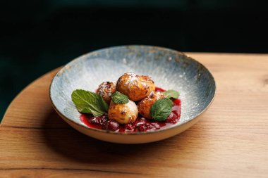 Tatlı hamur köfteleri taze nane yapraklarıyla süslenmiş ahşap bir masada parlak meyve sosunun içinde dinleniyor. Bu yemek lezzetli bir tat ve renk karışımı sergiliyor..