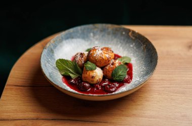 Hamur köfteleri böğürtlen sosundan bir yatağa konur ve nane yapraklarıyla süslenir. Tabak ahşap bir masada yuvarlak bir kasede sunuluyor..