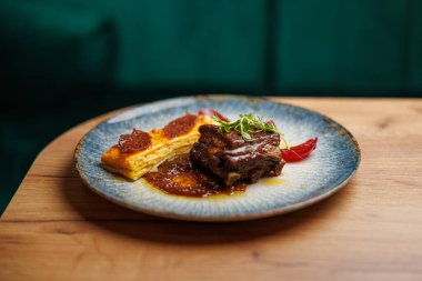 Etli ve yumurtalı muhallebi tabakları mavi bir tabakta sos ve garnitürle sunuluyor. Dekor, ahşap yüzeyler ve yeşil bir dekor içererek samimi bir yemek deneyimi yaratıyor..