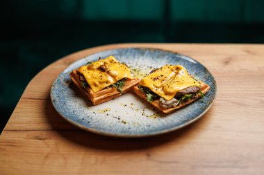 Erimiş peynir, yeşillik ve sığır eti içeren iki açık yüzlü sandviç dekoratif bir mavi tabağa yerleştirilir. Bu tabak ahşap bir masada sıradan bir yemek masasında servis edilir..