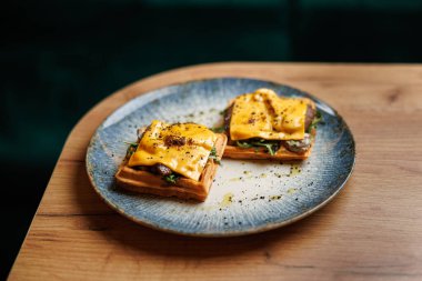 Erimiş peynir ve taze yeşilliklerle kaplı tuzlu waffle 'lar dekoratif bir tabakta servis edilir. Tabak, rahat bir restoranda, ahşap bir masanın üzerinde duruyor..