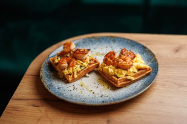 Waffles topped with scrambled eggs and grilled shrimp are presented on a blue plate, placed on a wooden table. This dish showcases flavors and textures together.