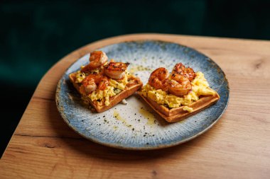 Shrimp are cooked and placed on top of scrambled eggs on two waffles. The dish is served on a decorative blue plate on a wooden table in a dining area.