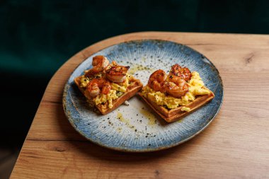 Shrimp cooked to perfection sit atop fluffy scrambled eggs and waffles, all plated on a stylish blue dish on a wooden table in an inviting space.