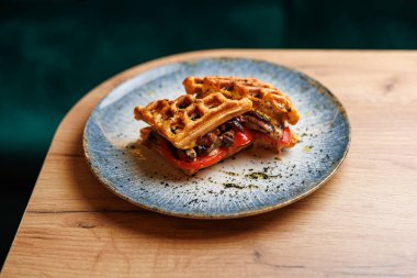 A waffle sandwich contains grilled vegetables and herbs, placed on a blue ceramic plate. This dish sits on a wooden table at a cafe, showing colorful food presentation.