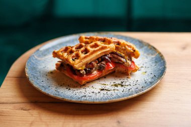 Grilled chicken and fresh tomatoes are layered between two waffles on a blue plate. This dish is served at a cafe during the afternoon. It invites you to savor each bite.