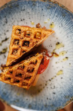 Two pieces of golden-brown waffles are served on a round dish with a slice of tomato and a drizzle of olive oil. This meal is enjoyed during a casual lunch setting.