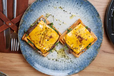 Two golden waffles sit on a blue plate, topped with melted cheese and greens, served at a casual dining spot during lunchtime, highlighting classic and modern flavors.