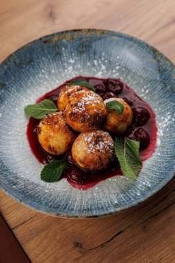 Hamur köfteleri kiraz soslu ve nane yapraklı bir kasede düzenlenir. Yemek, Ukrayna mutfağından geleneksel bir tarif sunarak lezzet ve dokuların karışımını vurguluyor..