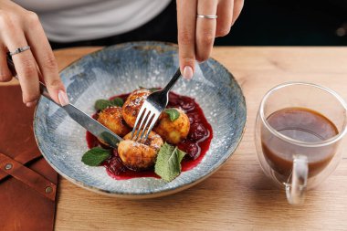 Bir insan Ukrayna hamur köftelerini böğürtlen soslu bir tabağa koymak için çatal ve bıçak kullanır. Bir fincan kahve yakınlarda, ahşap bir masada oturuyor..