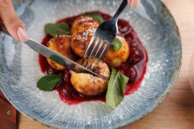 Elleri çatal ve bıçak ile pudra şekeri ile kaplanmış altın hamur işlerini kesmek için, canlı kiraz sosu ve taze nane ile çevrili. Sahne, aşçılık deneyimini yakalar..