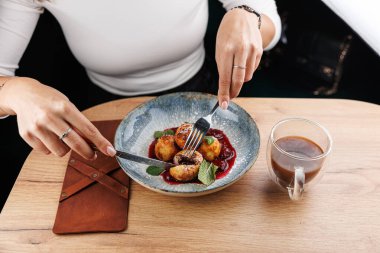 Kafe masasında bir tabak soslu Ukrayna hamur köftesiyle oturan bir kadın. Mantı kesmek için çatal ve bıçak kullanıyor. Yanında bir fincan kahve var..