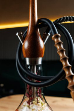 Bitkilerle dolu cam tabanlı rahatlatıcı bir nargile. Hortumlar ve ahşap bir gövde görülebilir olup, kullanıcıları aromalı tütünün tadını çıkarmaya davet ediyor.