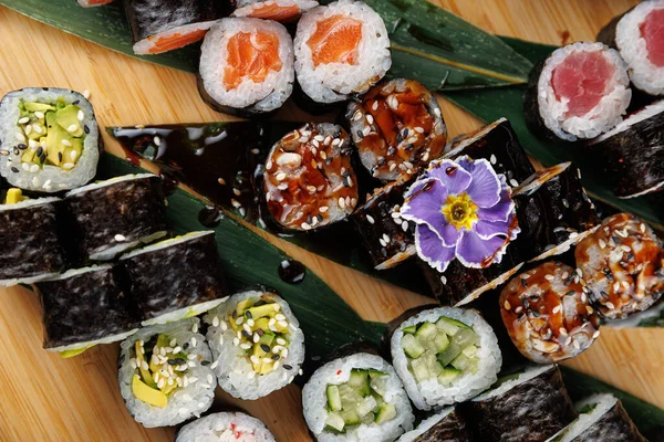 Suşi ruloları ahşap bir yüzeyde düzgün bir şekilde dizilir, somon ve avokado gibi farklı malzemeler sergilenir. Küçük çiçekler yemeğe dekoratif bir dokunuş ekler..